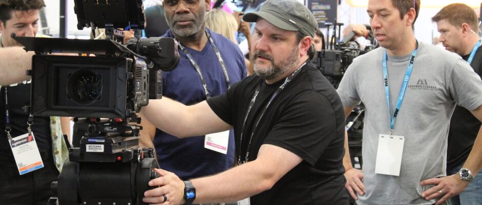A group of man crowd around a camera on a trade show floor.