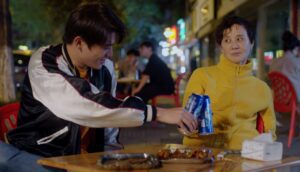 An older woman wearing a yellow sweater looks skeptically at a young man wearing a letterman jacket, they each hold blue beer cans and bring them together for a toast.