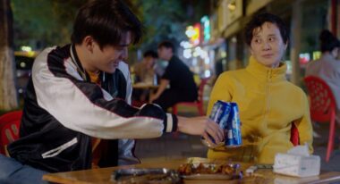 An older woman wearing a yellow sweater looks skeptically at a young man wearing a letterman jacket, they each hold blue beer cans and bring them together for a toast.