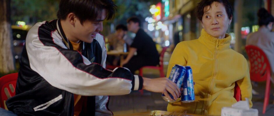 An older woman wearing a yellow sweater looks skeptically at a young man wearing a letterman jacket, they each hold blue beer cans and bring them together for a toast.