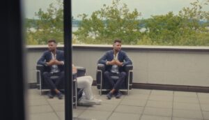A man in a business casual suit sits in a chair on an outside patio.