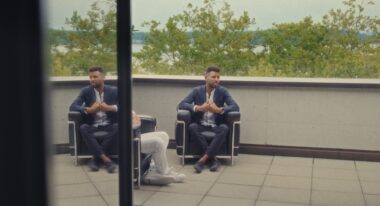 A man in a business casual suit sits in a chair on an outside patio.