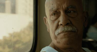 An older Latino man, who is bald and has a white mustache, looks out of a bus window.