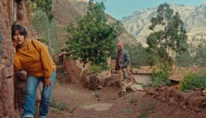 A young woman wears a yellow sweater and blue jeans, she leans a stone edifice and presses her ear against the wall.