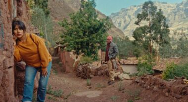 A young woman wears a yellow sweater and blue jeans, she leans a stone edifice and presses her ear against the wall.
