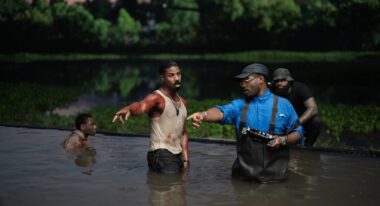 A man in a blood-stained undershirt and a man in overalls stand in a pool of water outdoors.