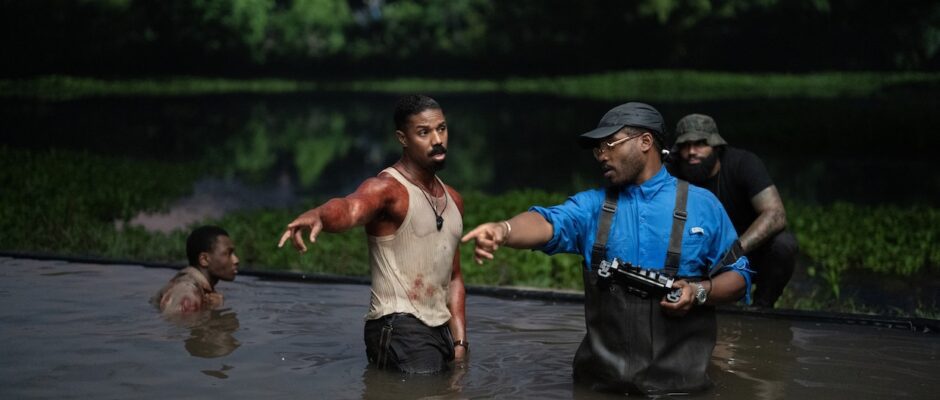 A man in a blood-stained undershirt and a man in overalls stand in a pool of water outdoors.