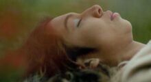 A close-up of the face of a Honduran teenage girl as she lays in the grass. She has long black hair and wears metallic blue eyeshadow.