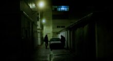 Two people on either side of a car in a dimly lit alley;
