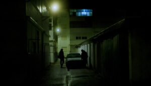 Two people on either side of a car in a dimly lit alley;