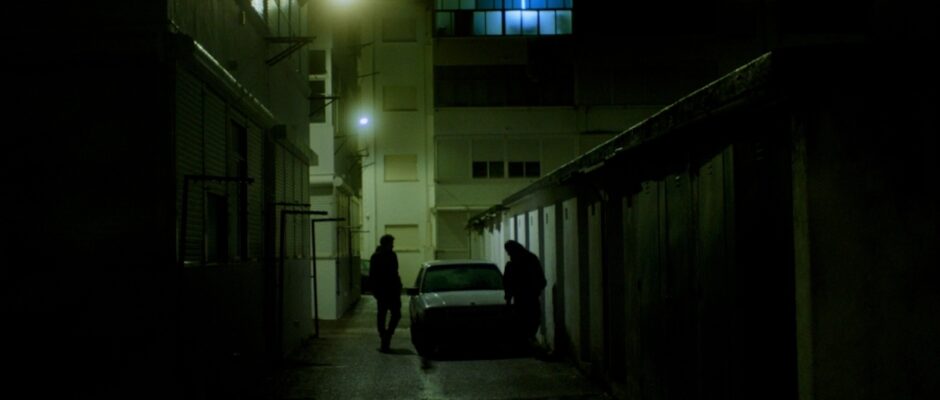 Two people on either side of a car in a dimly lit alley;