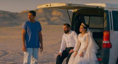 Three people, two of whom are seated on the back of a car, in the desert.