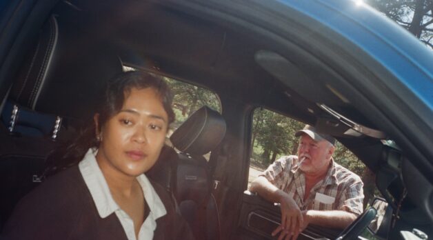 A woman sits in the right seat of a car as a man smoking a cigarette leans in through the left passenger window.