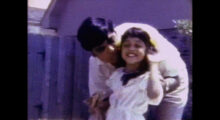 A still from a VHS home movie of an Indian father, with cropped black hair and a white shirt, bending down to kiss his young daughter on the head; she has long black hair and also wears white.
