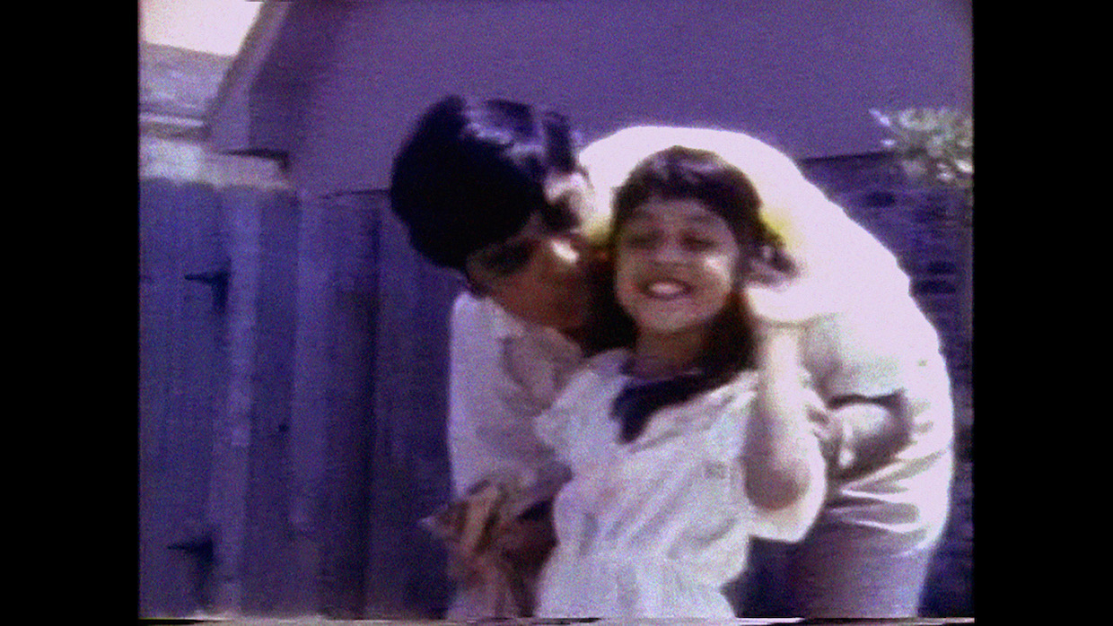 The Gas Station Attendant | Filmmaker Magazine A still from a VHS home movie of an Indian father, with cropped black hair and a white shirt, bending down to kiss his young daughter on the head; she has long black hair and also wears white.