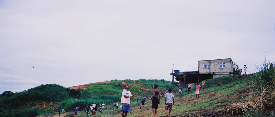 Children stand on a green hillside in Rio de Janeiro, they look up at the gray-blue sky, where they fly colorful paper kites.