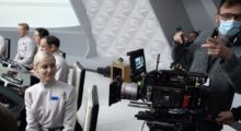 A cinematographer wearing a face mask stands nexts to a camera on a spaceship set.