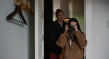A young woman with brown hair and a brown coat holds a camera to her face and takes a picture while standing in a doorway, a man with a maroon beanie looks over her shoulder.