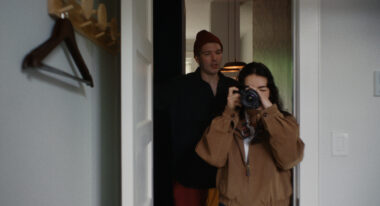 A young woman with brown hair and a brown coat holds a camera to her face and takes a picture while standing in a doorway, a man with a maroon beanie looks over her shoulder.