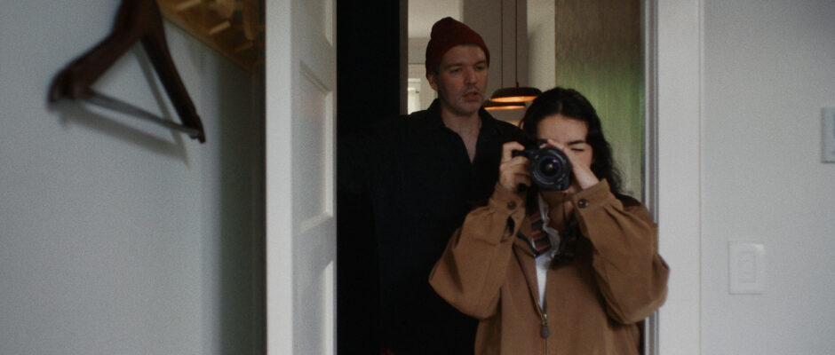 A young woman with brown hair and a brown coat holds a camera to her face and takes a picture while standing in a doorway, a man with a maroon beanie looks over her shoulder.