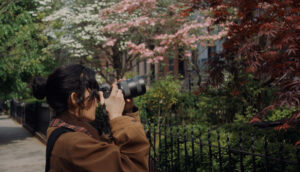 A young woman with her long dark hair tied back turns to the side and takes a picture of the front of a house, which is out of frame. She stands among trees that bloom with pink and white flowers.