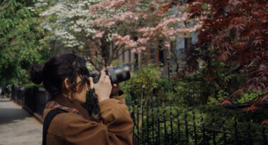 A young woman with her long dark hair tied back turns to the side and takes a picture of the front of a house, which is out of frame. She stands among trees that bloom with pink and white flowers.
