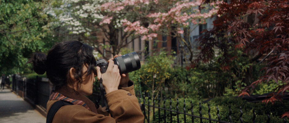 A young woman with her long dark hair tied back turns to the side and takes a picture of the front of a house, which is out of frame. She stands among trees that bloom with pink and white flowers.