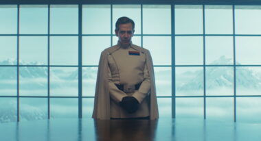A man in a white military suit and cape stands before a window facing out onto mountains.