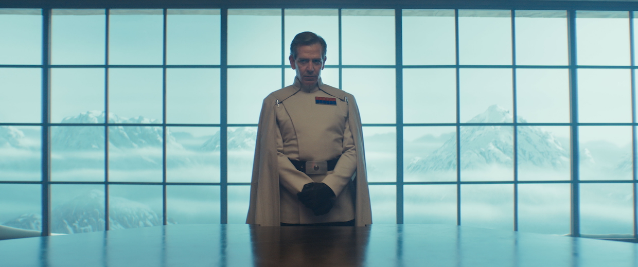 A man in a white military suit and cape stands before a window facing out onto mountains.