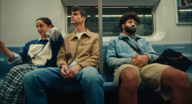 A young woman with brown hair, a tall man with salt and pepper hair and a man with a beard and afro sit together on a subway seat.
