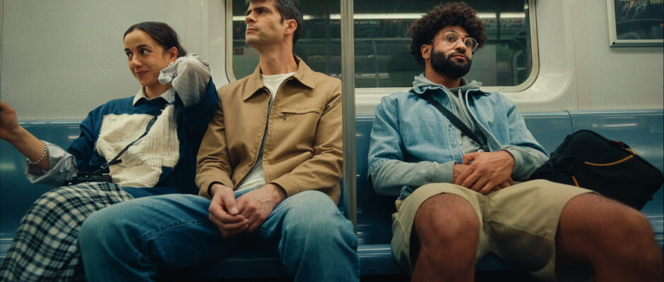 A young woman with brown hair, a tall man with salt and pepper hair and a man with a beard and afro sit together on a subway seat.