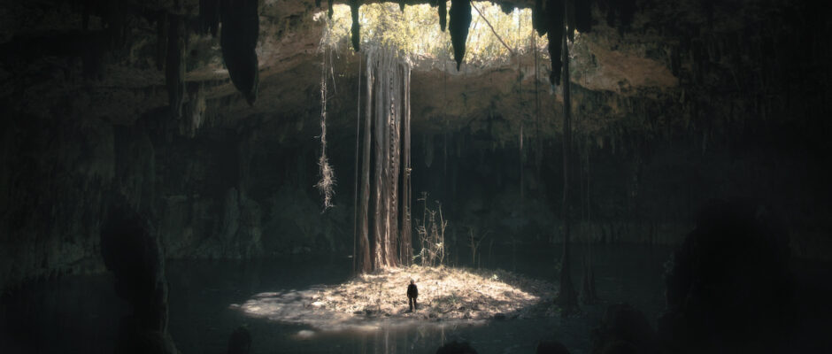 A beam of sunlight radiates town into a dark cavern, where a person stands and looks incredibly small amid the massive, dark space.
