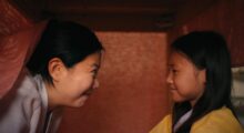 A mother and daughter in Traditional Korean dresses smile at each other as they sit under a dining table.