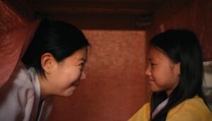 A mother and daughter in Traditional Korean dresses smile at each other as they sit under a dining table.