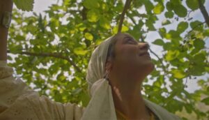A young woman wearing a white headscarf stands under a tree, she looks up to see the sky between the leaves.